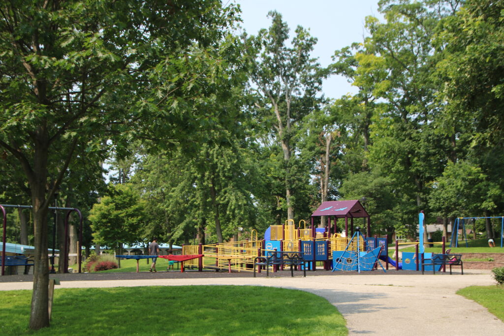 A photograph of a playground in a park. The playground is made of metal and plastic and has a slide, a climbing structure, a swing set, and a ramp. There are two people in the photo, one adult and one child. The adult is wearing a gray shirt and the child is wearing a red shirt. The playground is surrounded by trees and grass. The photo was taken on a sunny day.