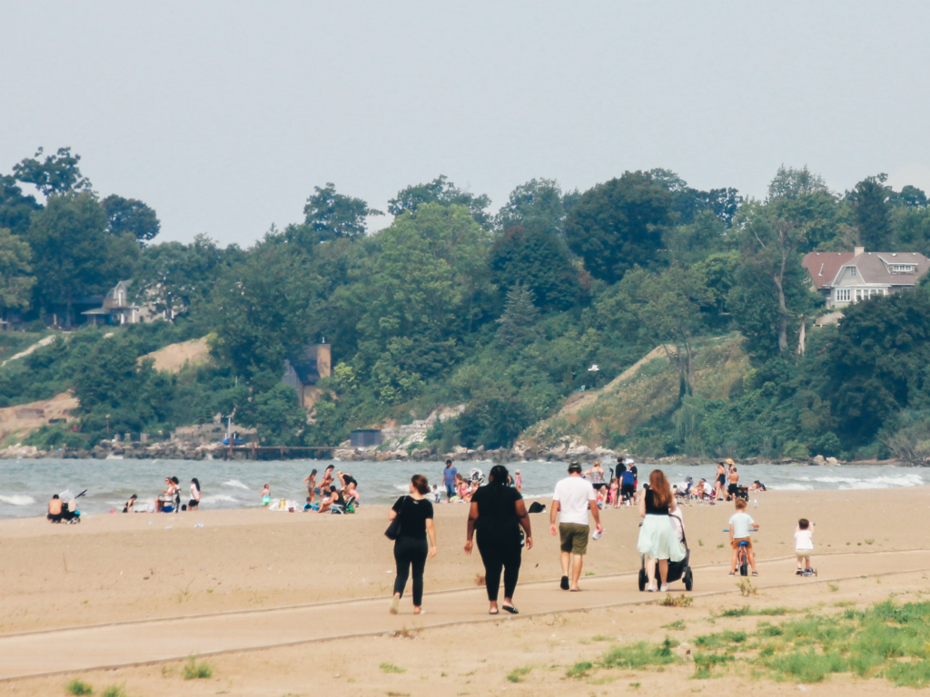 A photograph depicting a sandy beach scene with a group of people walking along a walkway. The path runs parallel to the shoreline. The background shows a line of trees and houses on a hillside overlooking the water. People are visible in the foreground and background, engaged in various activities such as walking, sitting, swimming and playing.