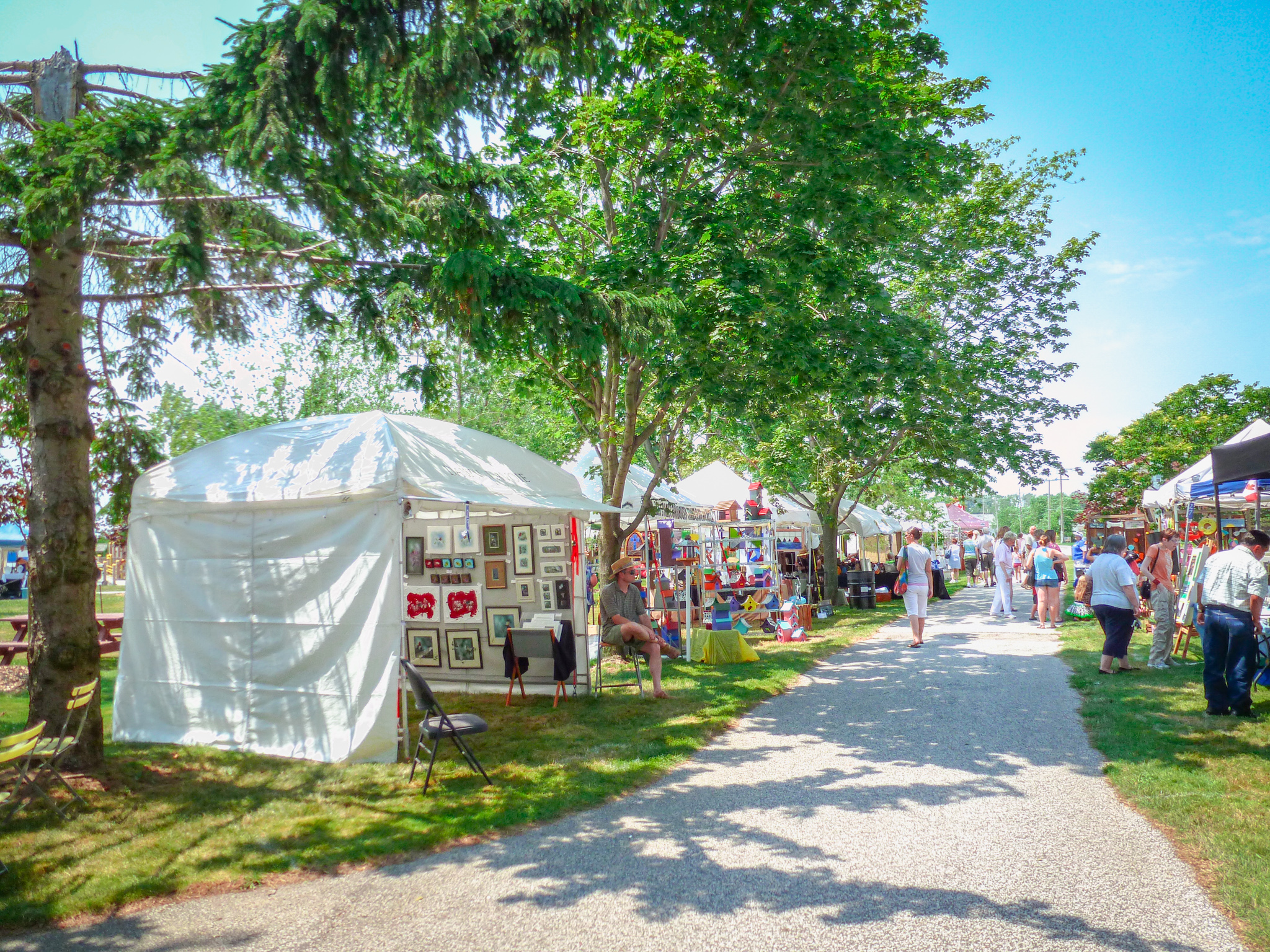 The image captures a vibrant outdoor art fair on a sunny day, with white tents lining a grassy path. Each tent showcases a variety of colorful artworks, including framed paintings, abstract sculptures, and handcrafted items. Visitors stroll leisurely, some stopping to admire the displays, while a few artists sit nearby, ready to engage with potential buyers. The scene is lively and bustling, with a mix of people enjoying the creative atmosphere under the shade of leafy trees.