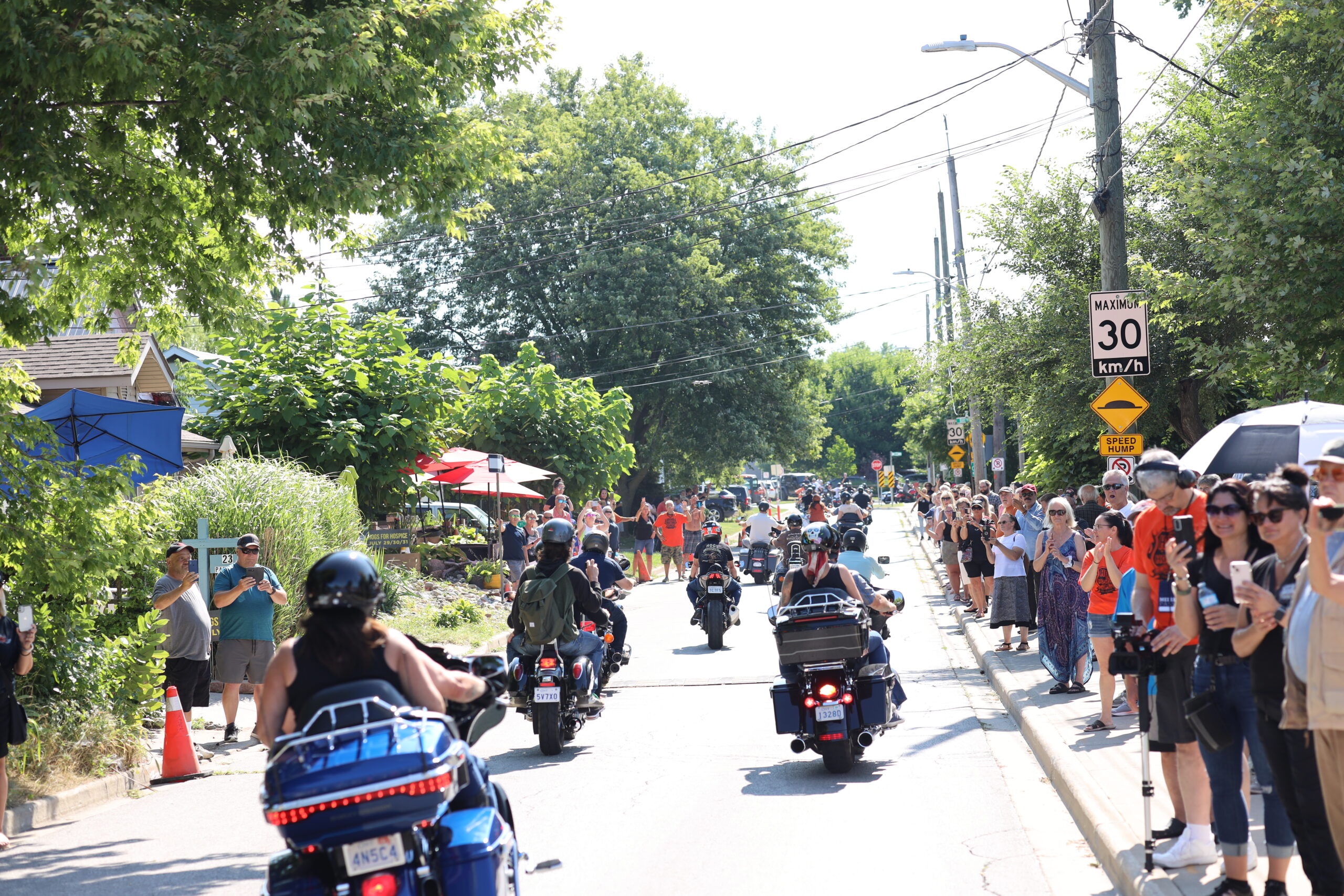 A photograph taken from a low angle, looking down a residential street lined with people on the right side of the frame. The people are standing on the sidewalk and are looking towards the left side of the frame. There are several motorcycles in the street moving in the same direction. The motorcycles are all black and chrome, and they have large saddlebags. The people on the motorcycles are wearing helmets and leather jackets. There are trees on both sides of the street, and the sky is blue. The road is paved and there is a speed limit sign on the right side of the frame. The sign is yellow with black lettering and reads "Maximum 30 km/h". There is also a sign below it that reads "Speed Hump". The photograph was taken on a sunny day.