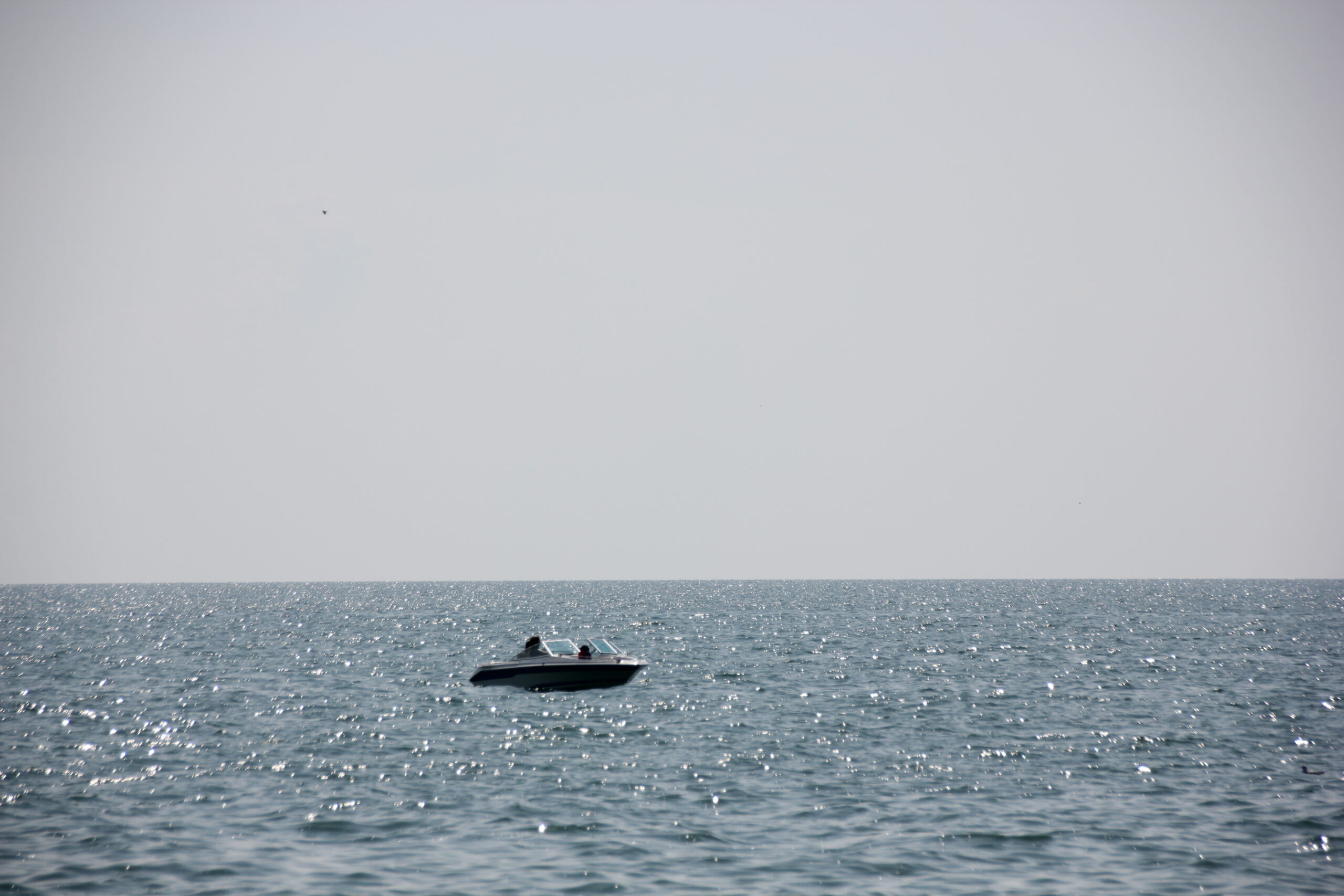 A photograph of a boat on the water. The boat is a small, white motorboat with a dark stripe running along the side. The boat is in the center of the image, and the water is a light blue color. The sky is a light blueish gray color.