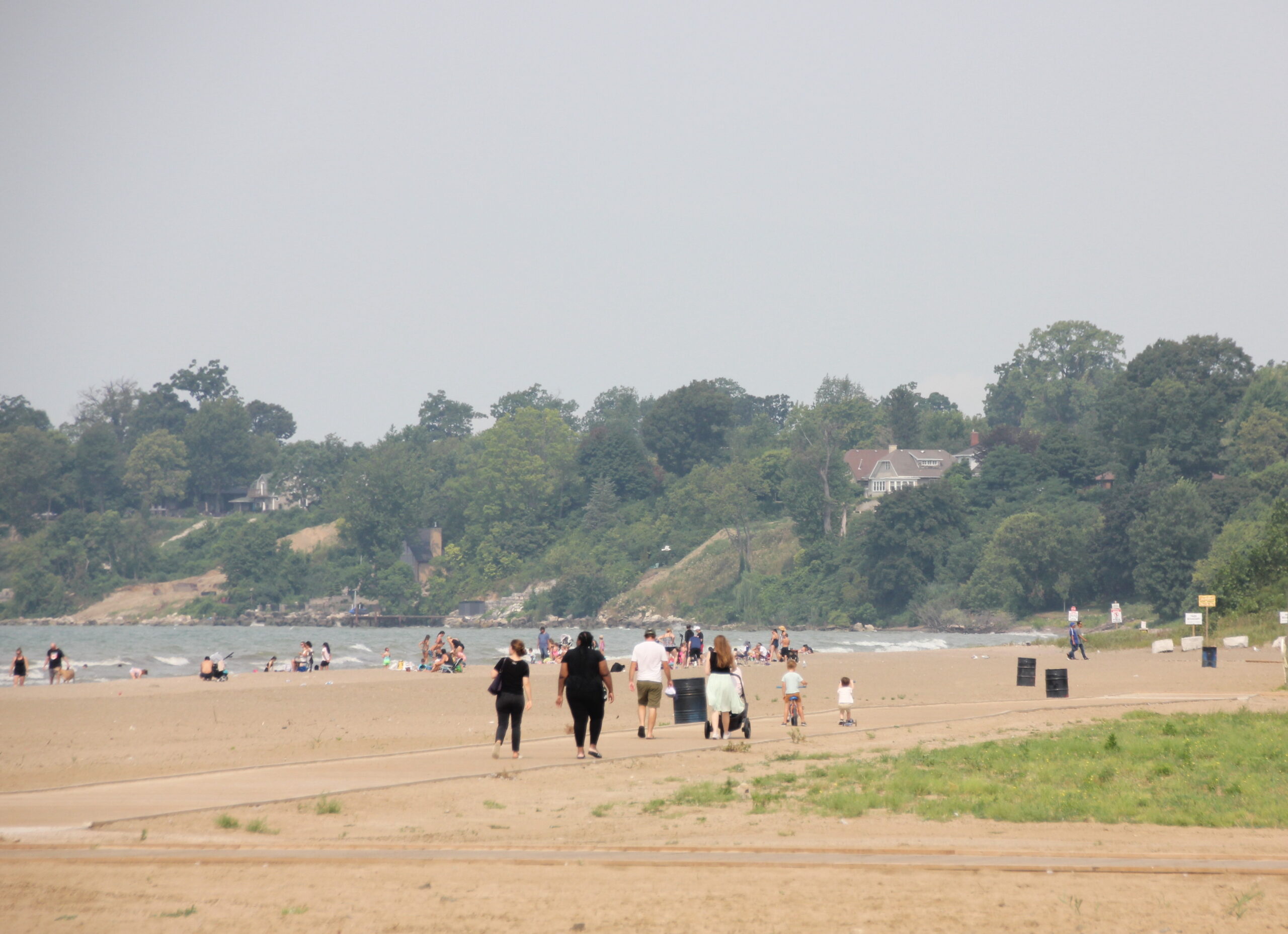 The subject is a group of people walking on a sandy beach. The background is a shoreline with trees and a house in the distance. The image is likely taken on a sunny day. The sand is a light tan color, and the water is a light blue color. The sky is a light blue color with some clouds. The trees are a dark green color. The people are wearing casual clothing. The image is composed in a way that emphasizes the vastness of the beach and the open space.