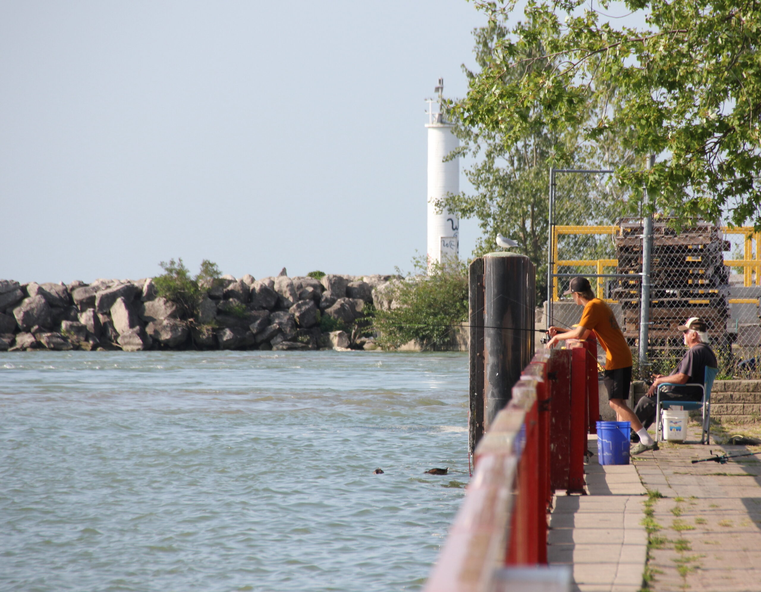 The image shows two people fishing from a pier. The pier is made of wood and has a red railing. The people are standing on the pier and are looking out at the water. There is a lighthouse in the background. The water is choppy and there are some rocks in the foreground. The sky is blue and there are some clouds in the distance.