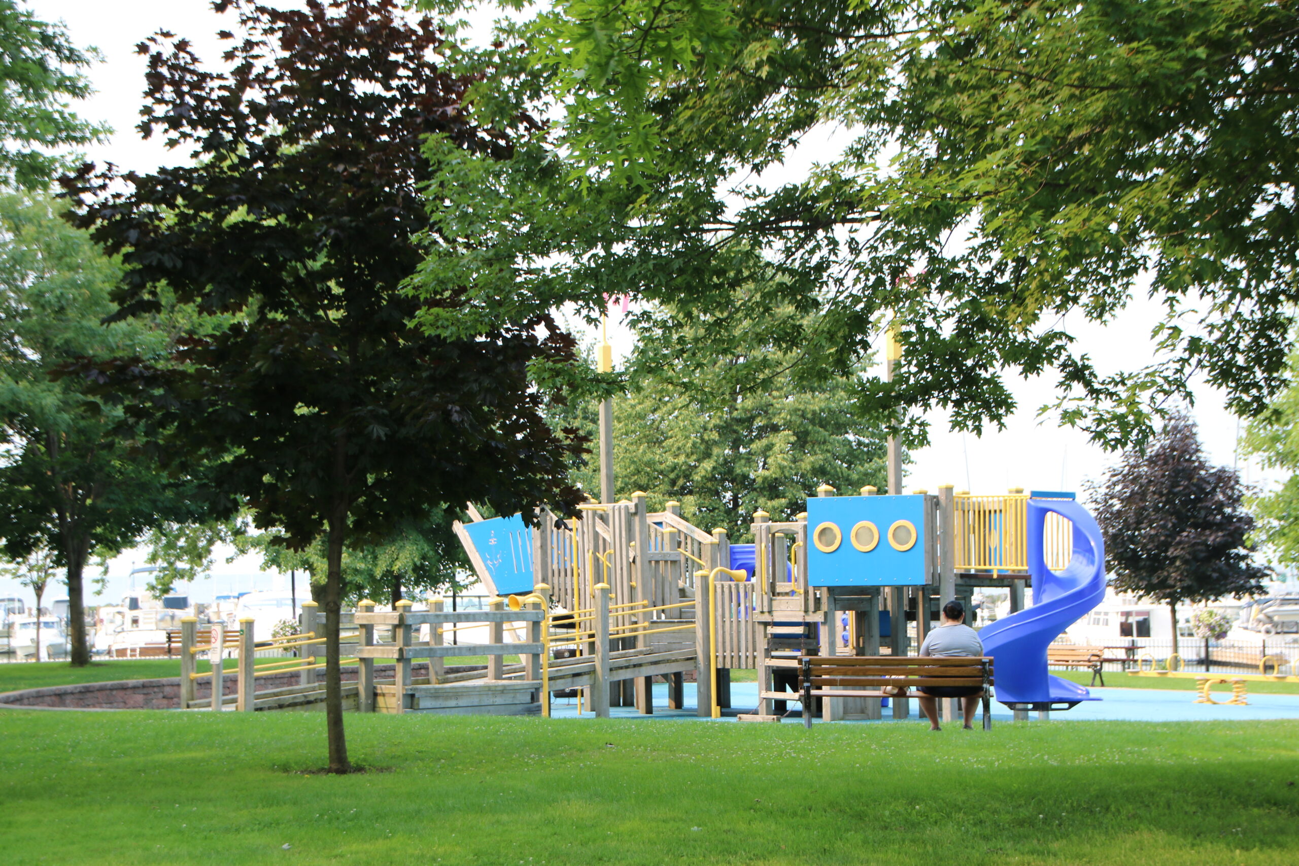 In this serene park scene, a vibrant playground stands amidst lush greenery, its structures painted in cheerful blues and yellows. The playground features a slide and various climbing elements, inviting children to explore and play. A solitary figure sits on a wooden bench, perhaps enjoying the tranquility and watching over the area. The surrounding trees, with their dense foliage, provide a natural canopy, casting gentle shadows on the well-maintained grass. The overall atmosphere is peaceful and inviting, capturing a moment of quiet leisure in a community space.