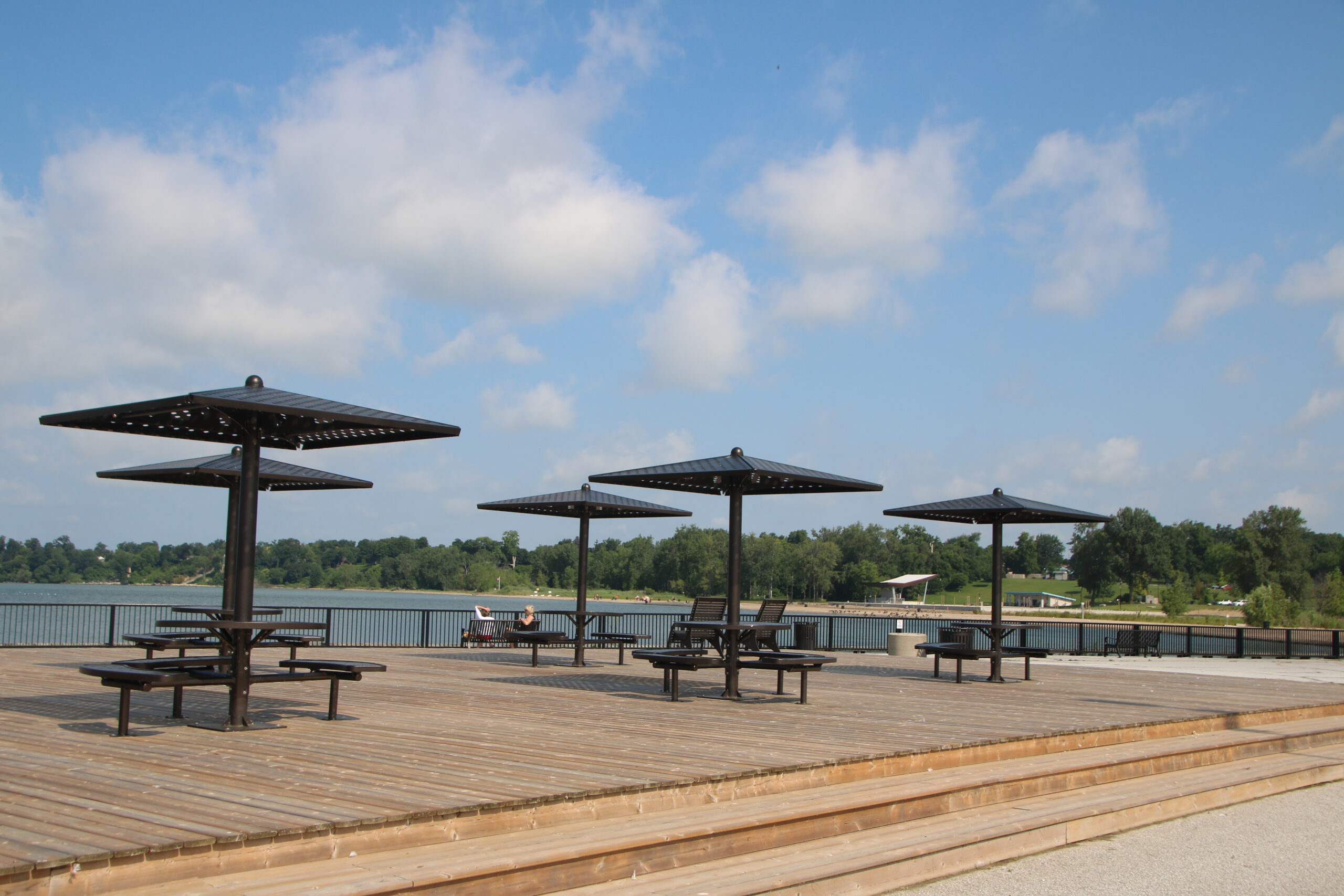 A photograph of a wooden deck with four black metal picnic tables with black metal umbrellas. The deck is made of wood planks and has a railing along the edge. There are trees in the background and a body of water in the distance. The sky is blue with white clouds.