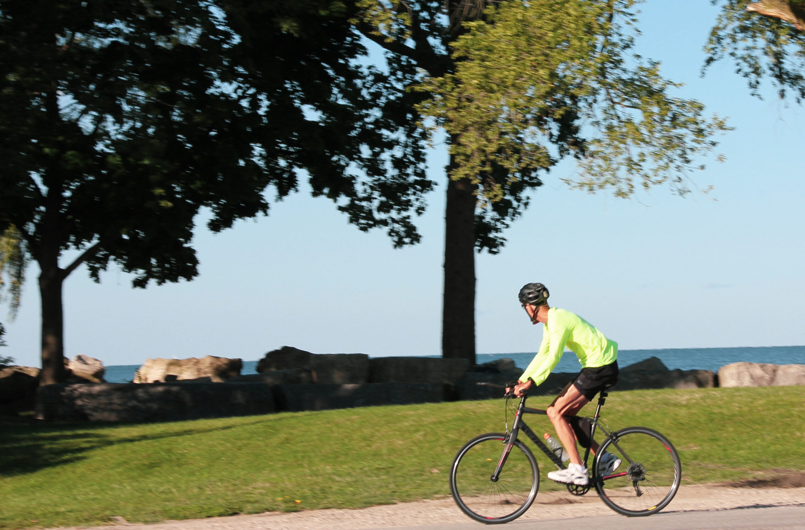 A photograph of a man riding a bicycle on a paved path. The man is wearing a neon green long-sleeved shirt, black shorts, and a black helmet. The bicycle is black with thin tires. The man is riding on a paved path with a grassy area to the right and a stone wall to the left. The background is a blurred image of trees and a body of water.