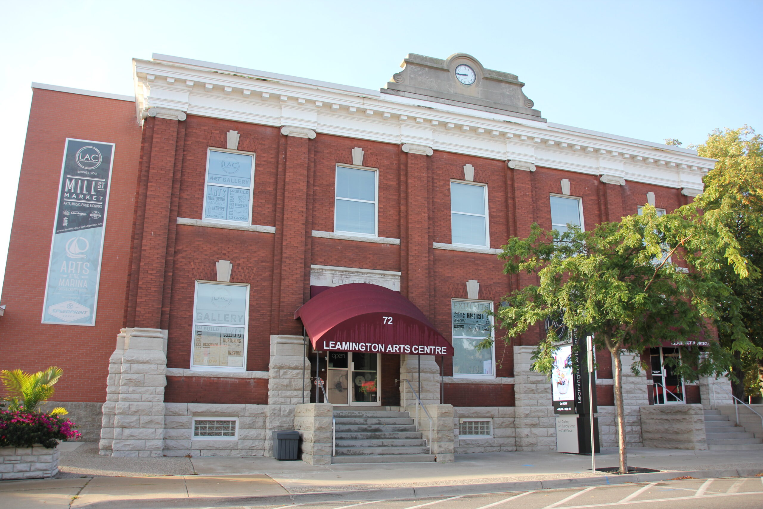 A photograph of a brick building with a white trim and a maroon awning over the entrance. The building has a clock tower on the roof. The building has several windows, some of which are covered with posters. The building is in front of a sidewalk and a street. There is a tree in front of the building. The building is a two-story building with a large sign that says "Leamington Arts Centre" above the entrance. The entrance is a set of steps leading up to a door. The door is open and there is a sign that says "open" on it. There is a sign on the side of the building that says "Art Gallery" and another sign that says "Arts Marina." There is a sign on the sidewalk in front of the building that says "Leamington Arts Centre."