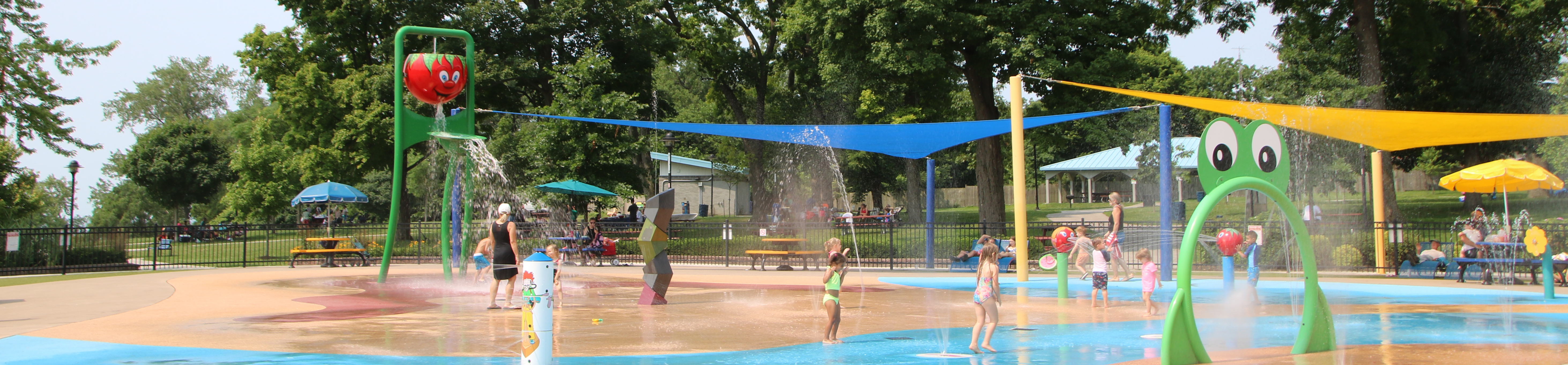 The image captures a lively splash pad in a park, bustling with children playing in the water. Brightly colored structures, including a green frog-shaped arch and a red bucket, spray water, creating a joyful atmosphere. The ground is a mix of blue and tan surfaces, mimicking water and sand. Overhead, vibrant blue and yellow shade sails provide relief from the sun. In the background, lush green trees frame the scene, adding a natural backdrop to this playful setting. Families relax under umbrellas, enjoying the sunny day.