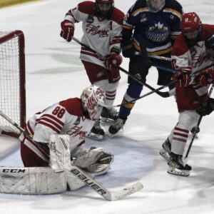 A live action shot from an ice hockey game showing players in red and white uniforms, with a goalie in the foreground wearing a goalie mask and holding a hockey stick. The goalie is positioned in front of a net with the CCM logo on it, while other players are seen on the ice, indicating a game in progress.