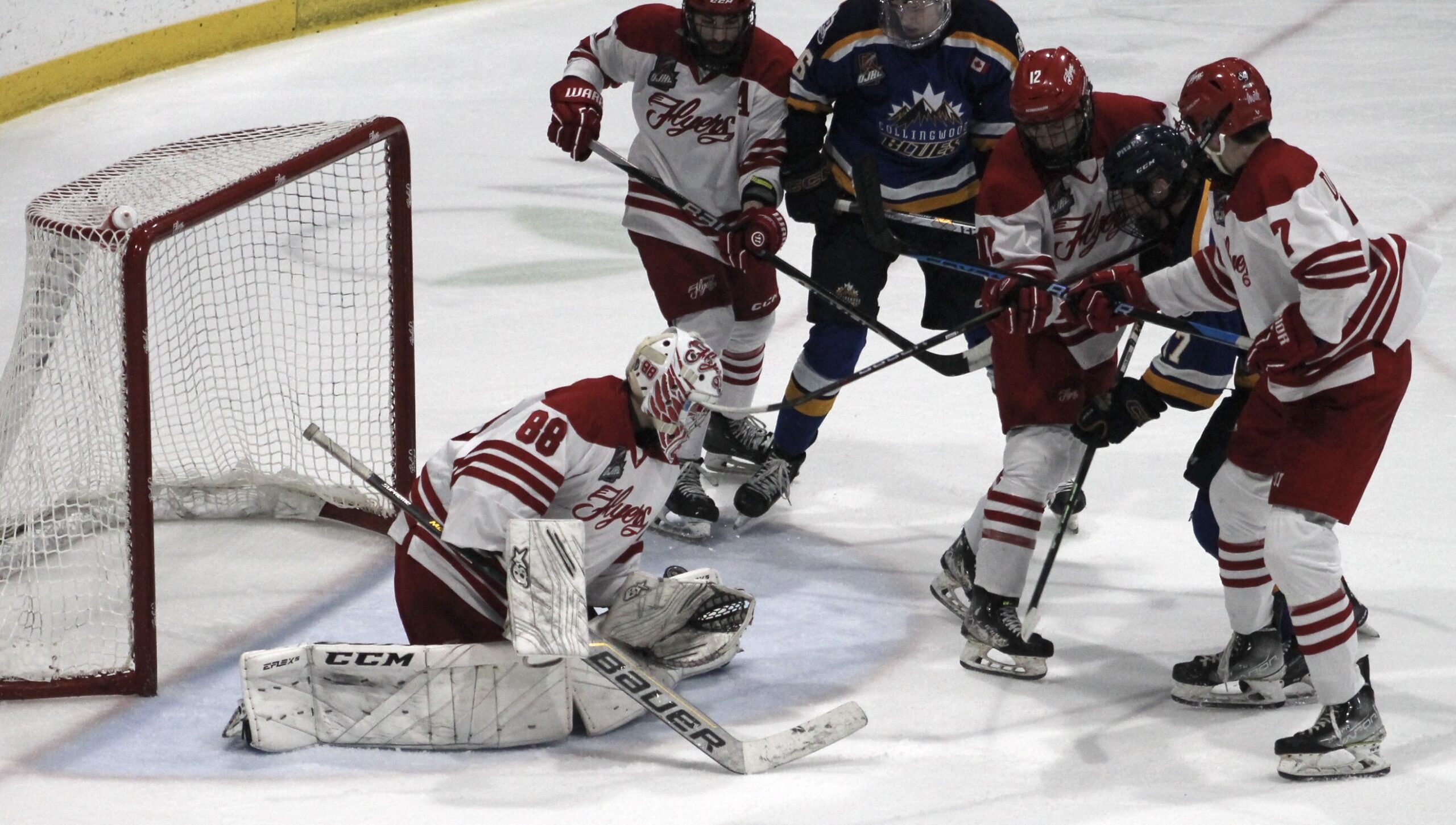 A live action shot from an ice hockey game showing players in red and white uniforms, with a goalie in the foreground wearing a goalie mask and holding a hockey stick. The goalie is positioned in front of a net with the CCM logo on it, while other players are seen on the ice, indicating a game in progress.