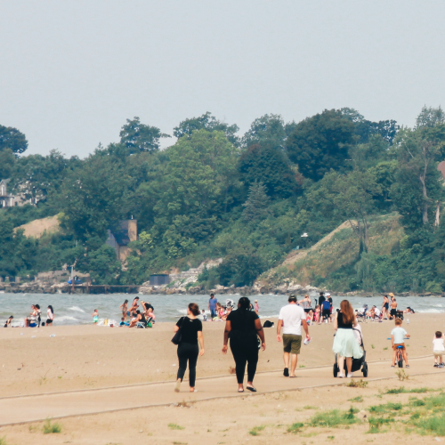 A photograph depicting a sandy beach scene with a group of people walking along a walkway. The path runs parallel to the shoreline. The background shows a line of trees and houses on a hillside overlooking the water. People are visible in the foreground and background, engaged in various activities such as walking, sitting, swimming and playing.